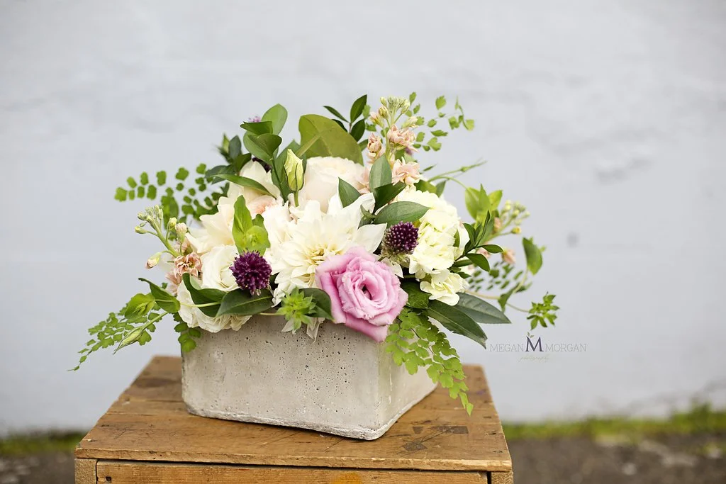 A flower arrangement with white, pink, and purple flowers in a rectangular concrete vase placed on a wooden surface against a neutral background.
