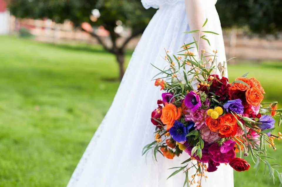 A bridal bouquet with orange roses, purple anemones, pink dahlias, and other colorful flowers, held by a bride dressed in a white wedding gown outdoors.