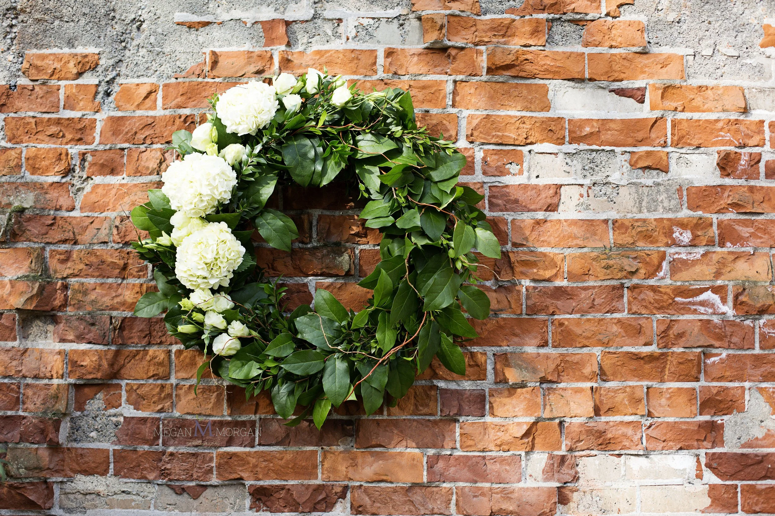 White floral wreath with greenery hanging on a red brick wall.