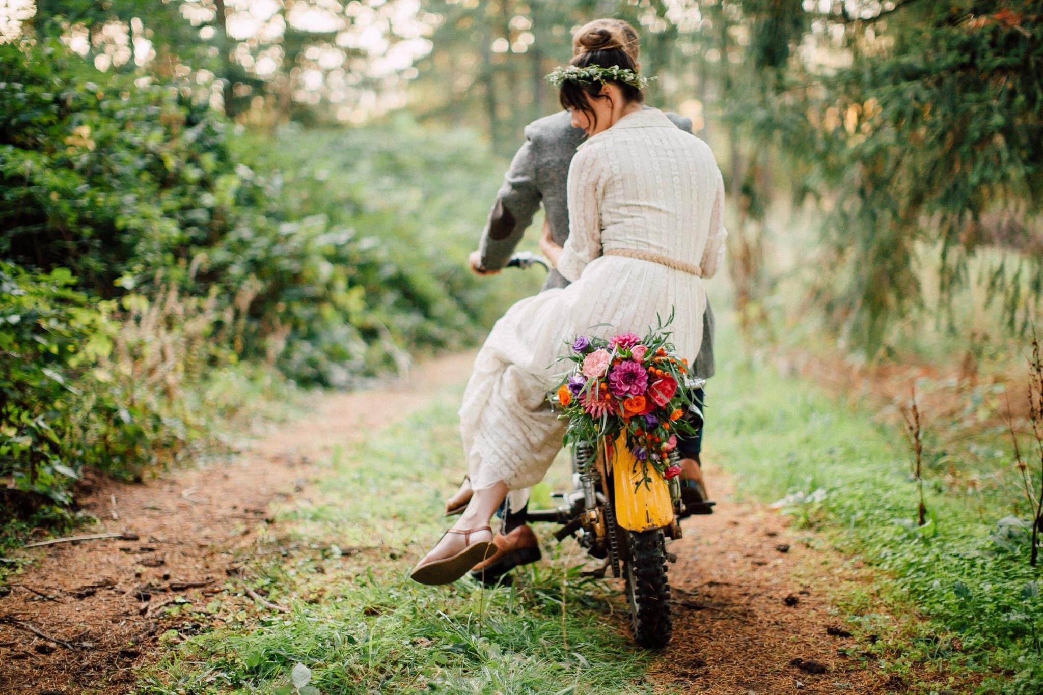 A woman in a white dress with a floral crown riding a dirt bike with a large colorful flower arrangement on the front through a forest path.