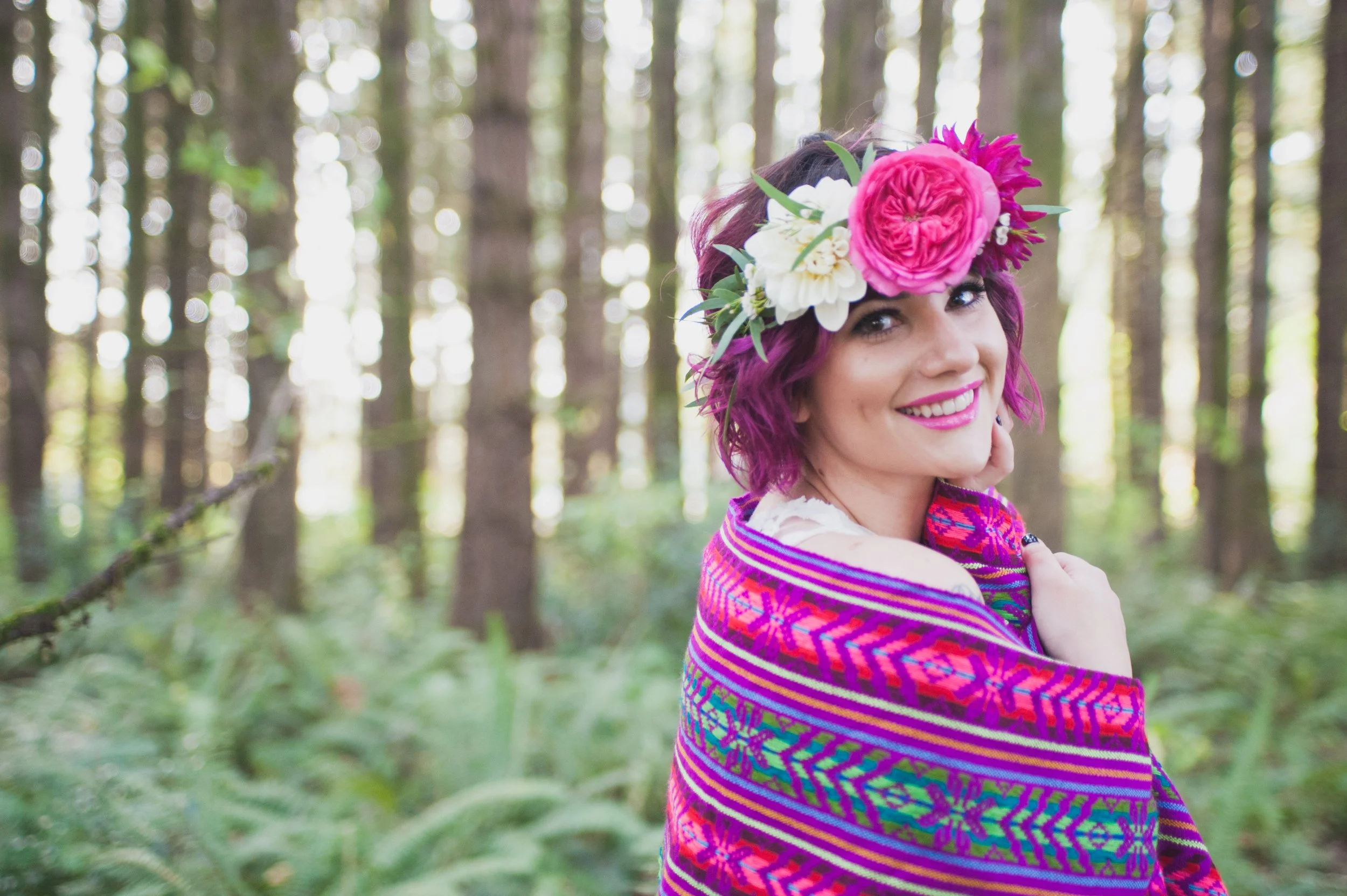 A woman with short purple hair and bright pink lipstick smiling in a forest, wearing a colorful shawl and a large flower crown with pink and white flowers.