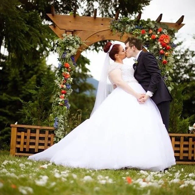A bride and groom share a kiss at an outdoor wedding ceremony, standing under a wooden arch decorated with colorful flowers, with trees and a cloudy sky in the background.