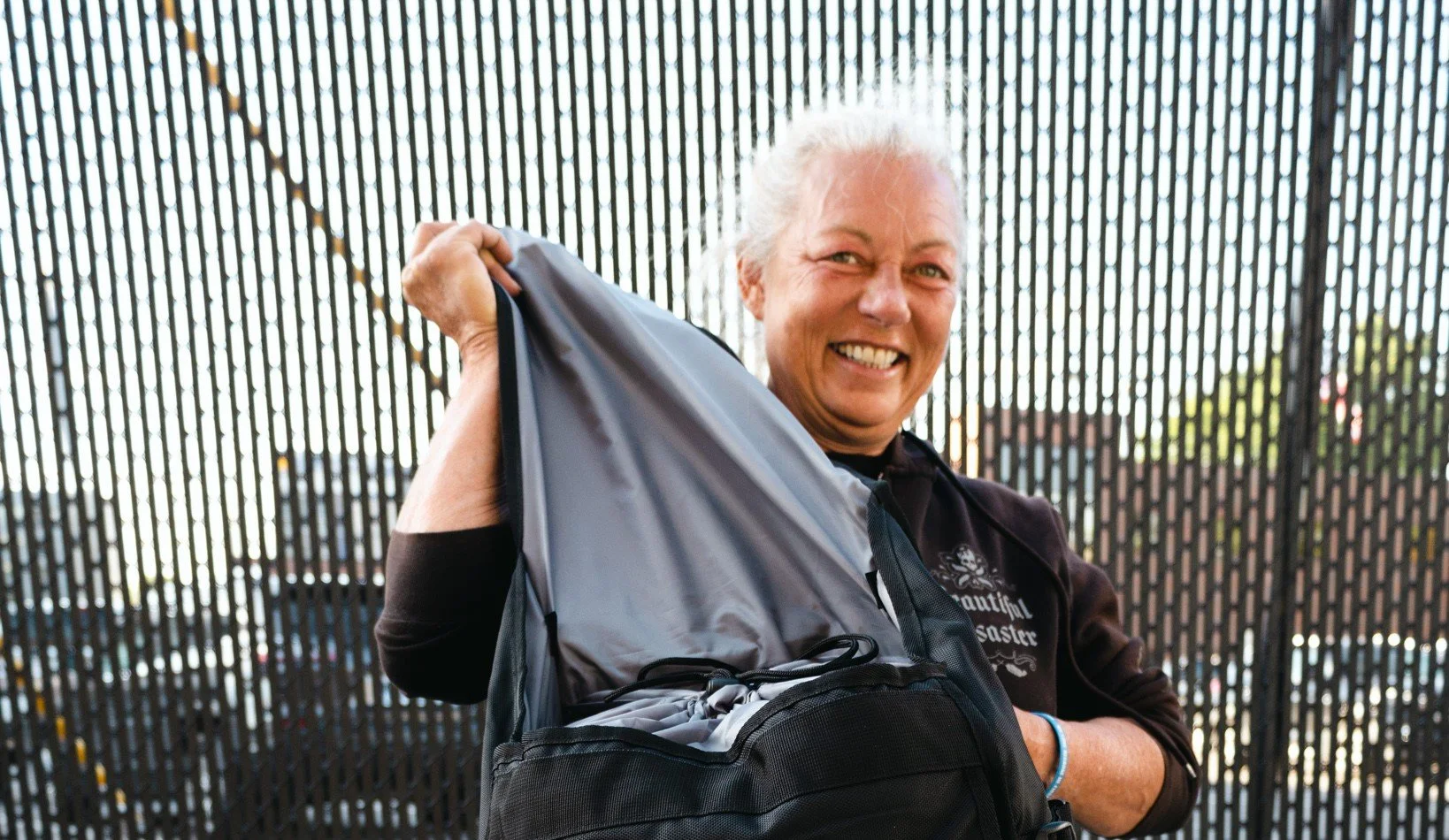 Smiling woman with gray hair in a black jacket holding a gray backpack in front of a black metal fence.