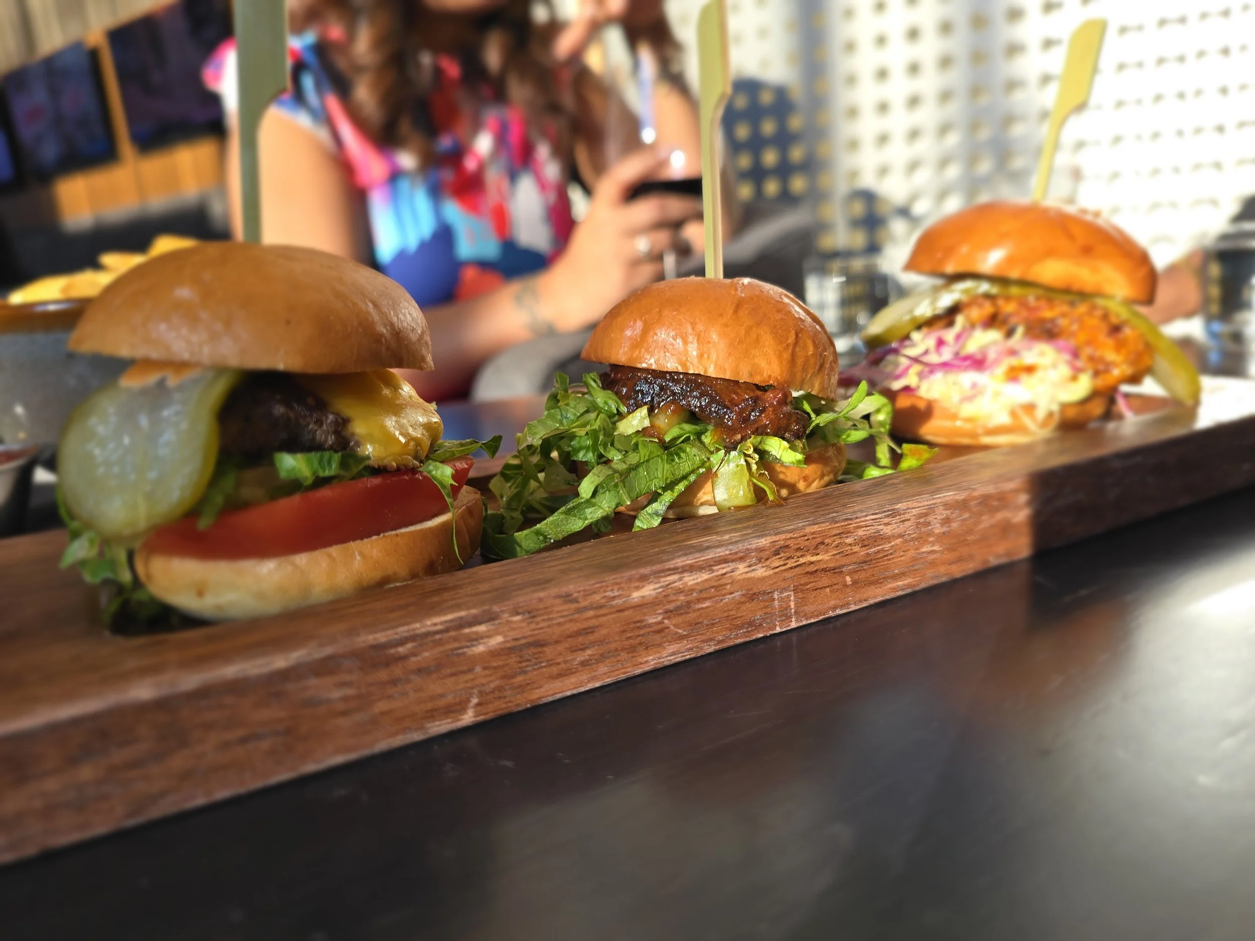 Three burgers on a wooden serving board in a restaurant setting with a person in the background using a phone.