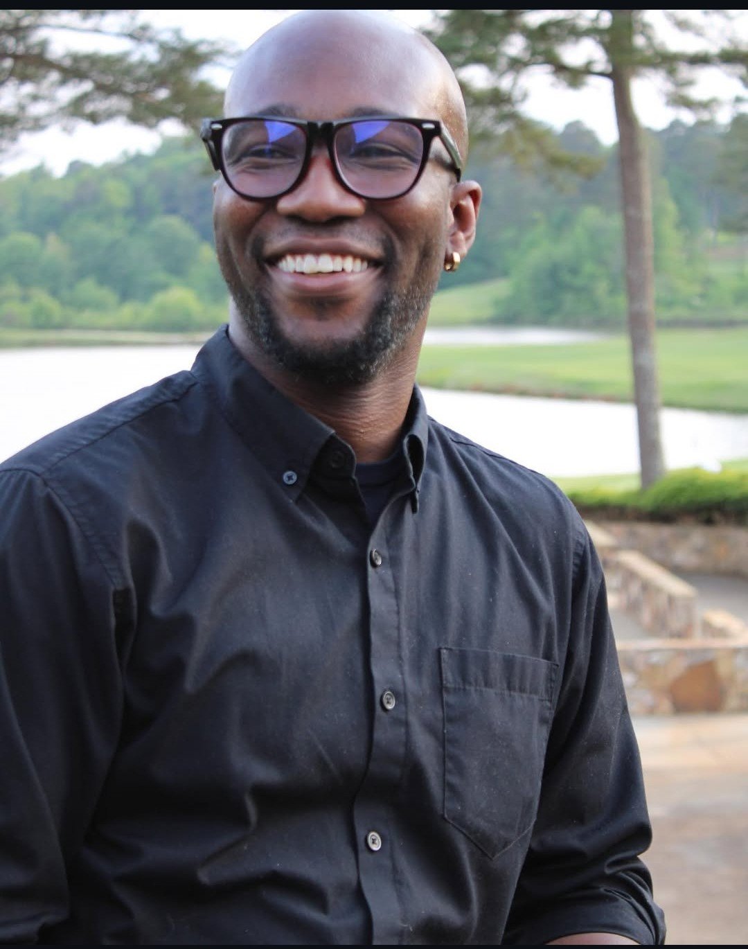 A smiling bald man with glasses and a gold earring, wearing a black button-up shirt, standing outdoors near a lake with greenery and trees in the background.