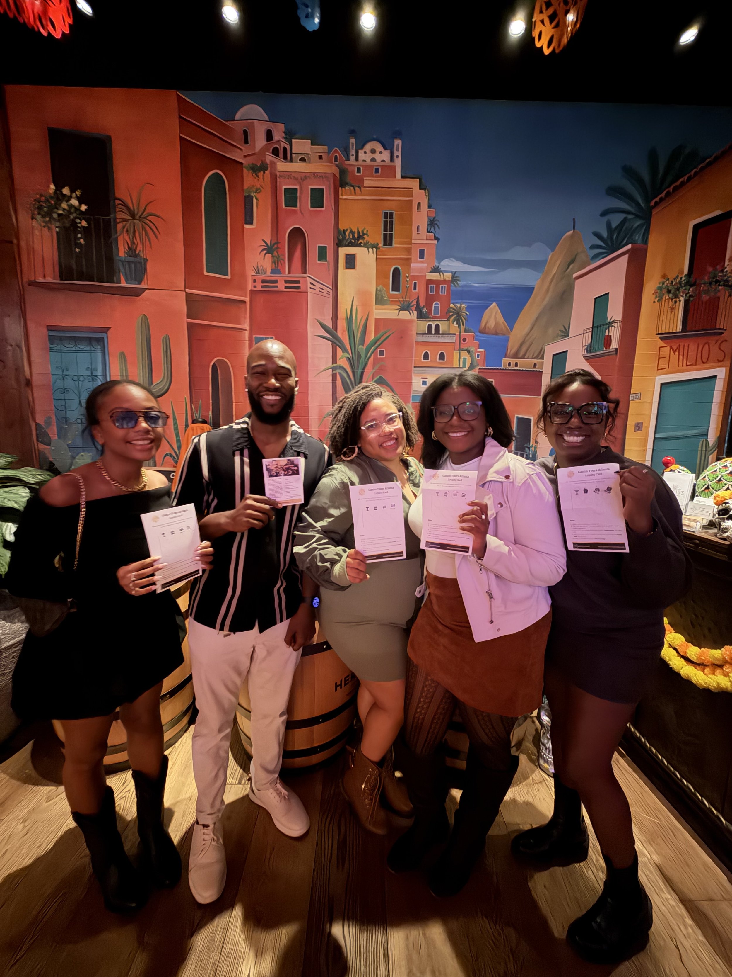 Five people standing together in front of a colorful mural, holding papers, smiling. The mural depicts a vibrant, seaside village scene with colorful buildings, palm trees, cacti, and mountains.
