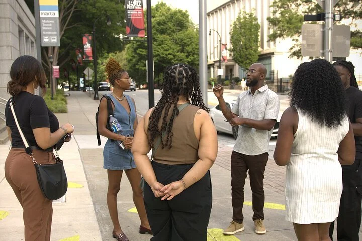 A group of six diverse people engaging in conversation on a city sidewalk with buildings and trees in the background.