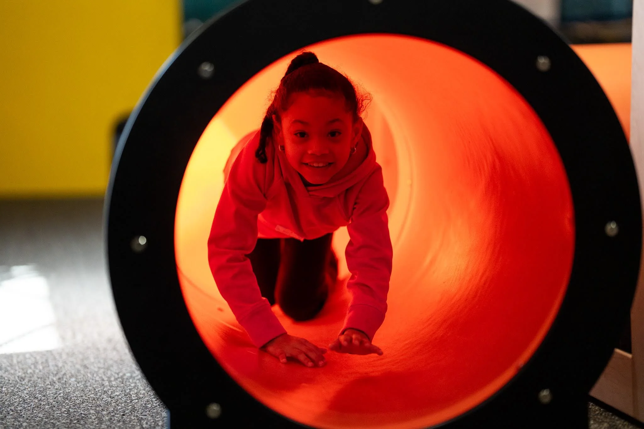 Child crawling through a brightly lit orange tunnel at an indoor play area.