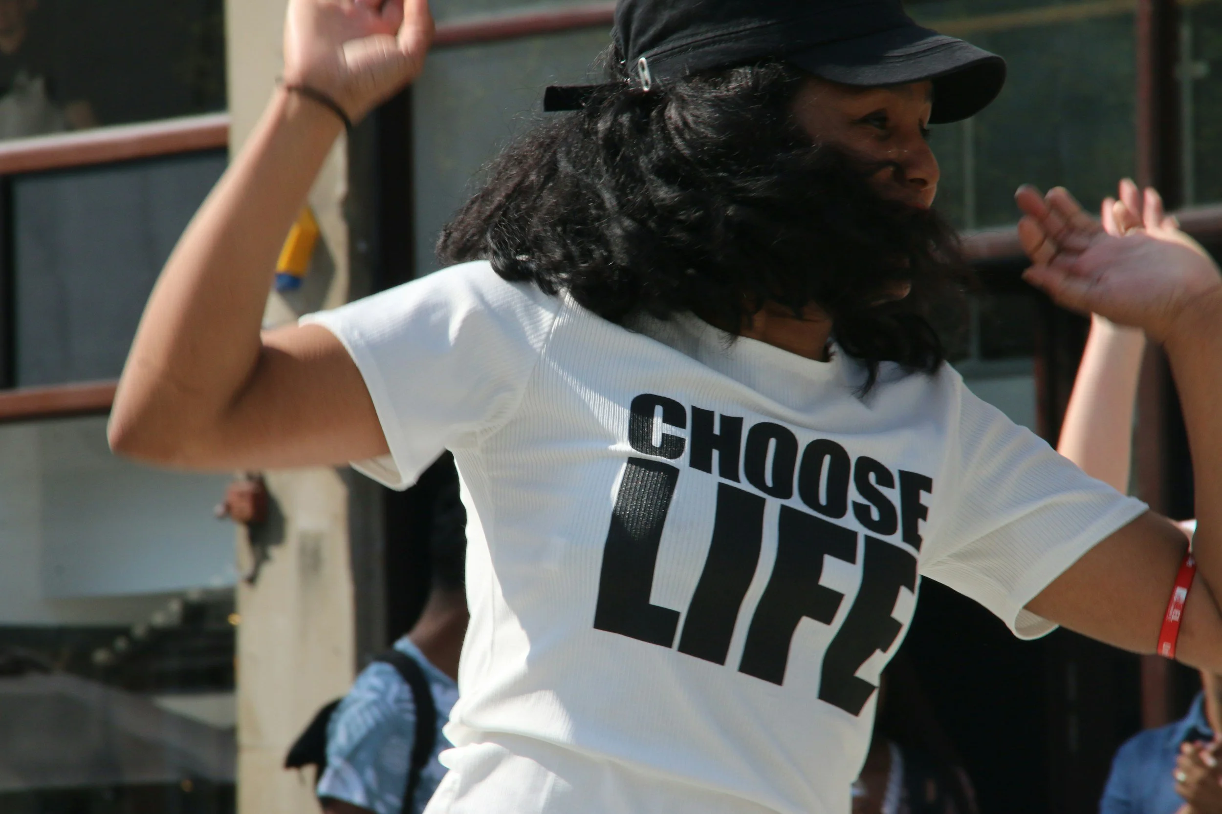 Person with long curly hair wearing a black cap and a white t-shirt that says 'CHOOSE LIFE', raising one arm in a fist and the other hand in a relaxed position, outdoors with other people visible in the background.