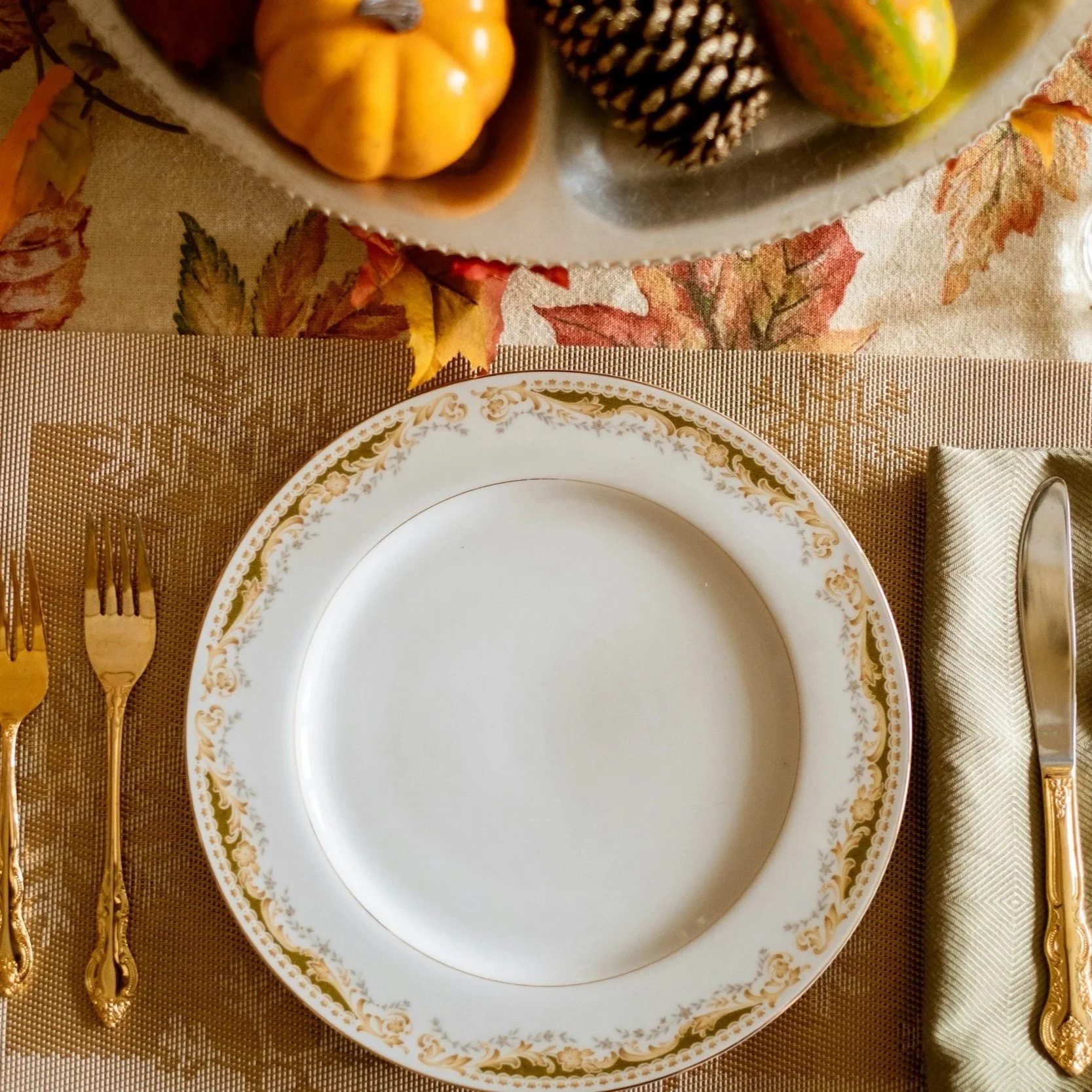 Empty white plate with gold floral border on a beige tablecloth, with gold utensils to the left and a rolled beige napkin with gold and silver utensils to the right, with a Thanksgiving-themed centerpiece of pumpkins, pinecones, and gourds on a table with an autumn leaf patterned tablecloth.