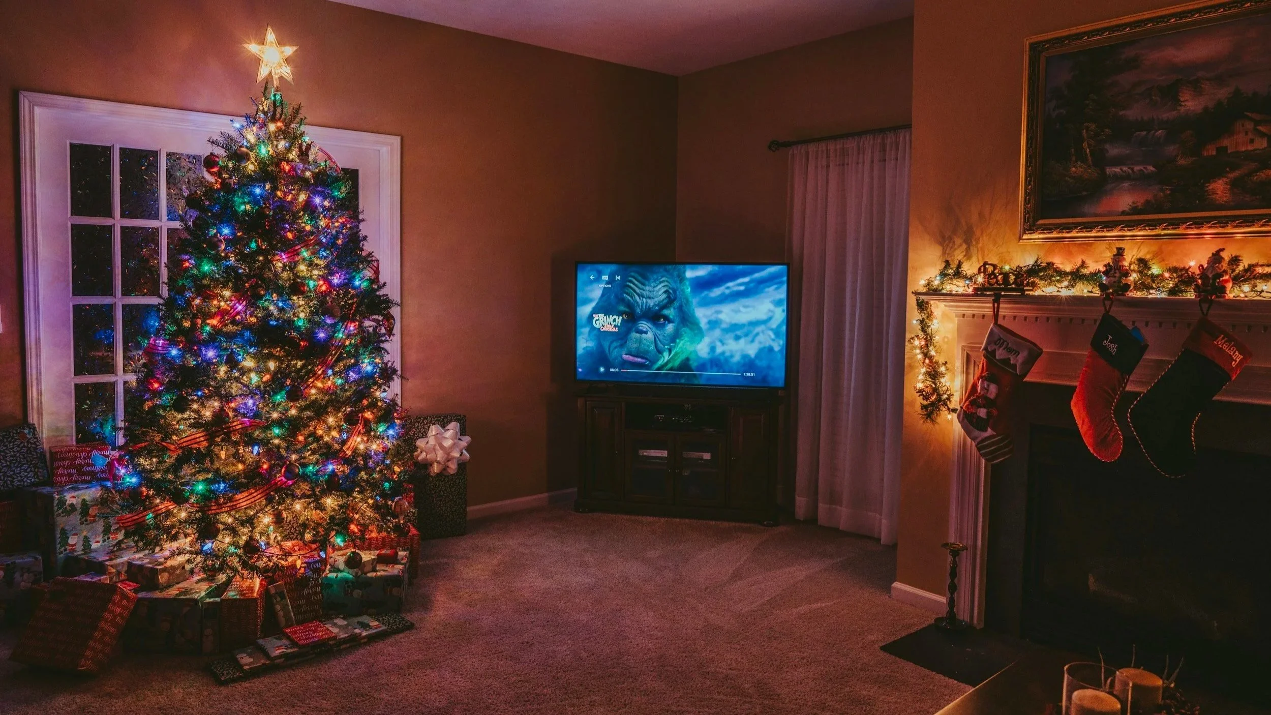 A decorated Christmas tree with multicolored lights and a star topper, surrounded by wrapped presents, stands in front of a window. To the right, a television shows a scene with a giant creature, and a lit fireplace decorated with holiday stockings and garland with ornaments hangs on the wall.