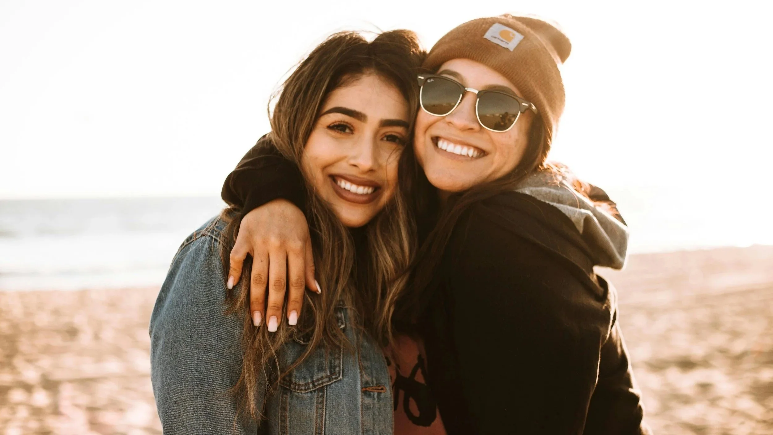 Two young women hugging on a beach at sunset, smiling and enjoying their time together.