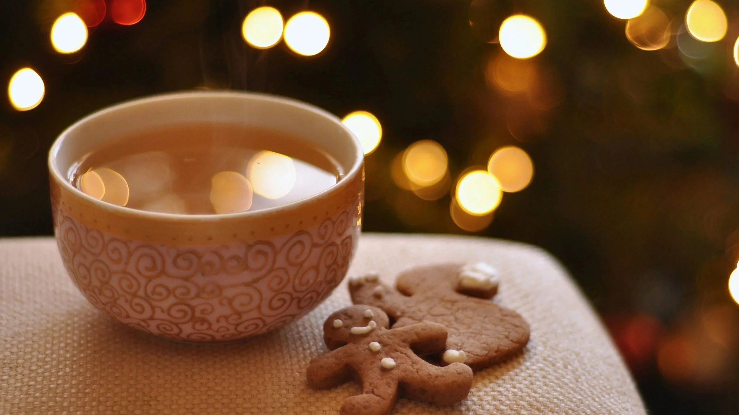 A cup of tea or hot chocolate on a white cloth surface, with gingerbread cookies shaped like a gingerbread man and a snowman beside it. The background features blurred warm yellow Christmas lights.