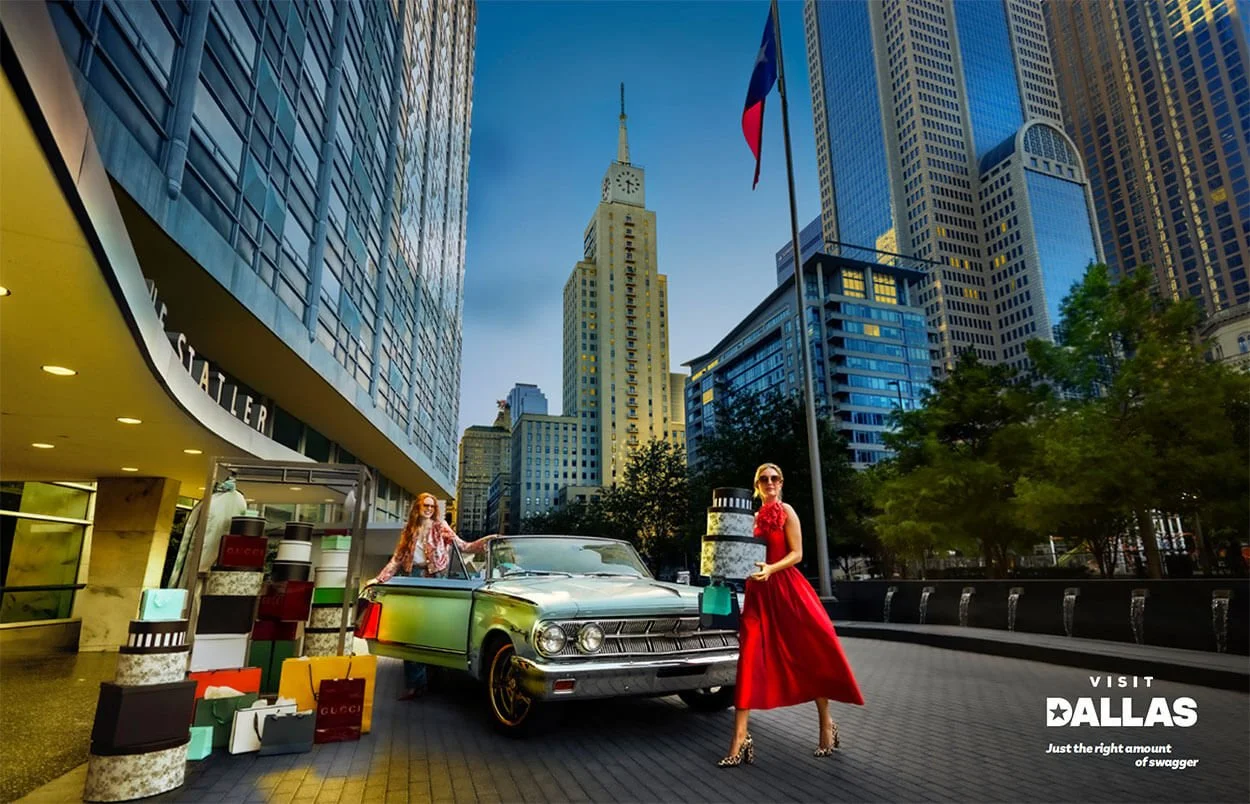 Two women with shopping bags posing on a city street in Dallas, with tall skyscrapers and a flag in the background. One woman is wearing a red dress and high heels, standing beside a vintage car, while the other is wearing a patterned blazer and sung