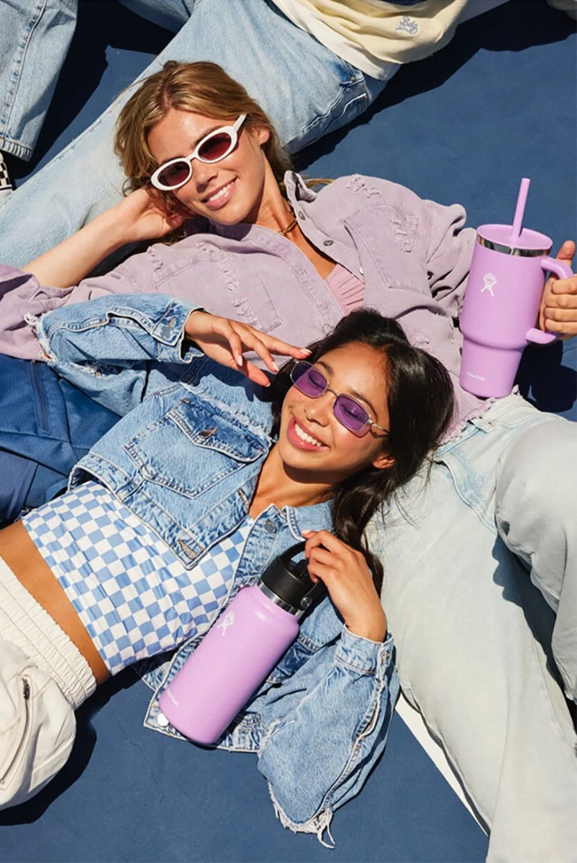 Two women lying on the ground, smiling, wearing sunglasses, holding purple Hydro Flask water bottles, and relaxing outdoors.