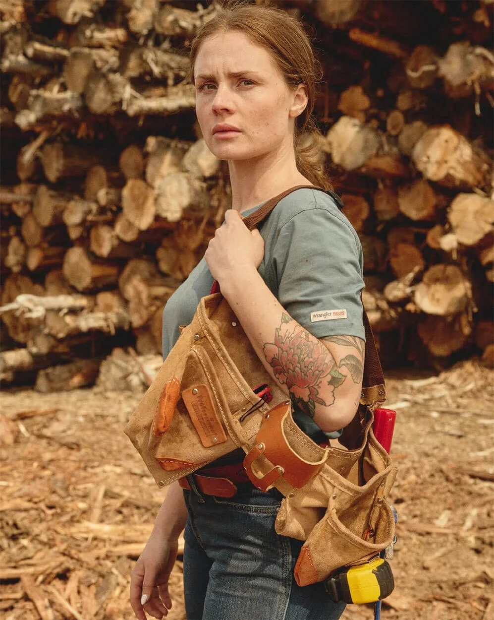 A woman with red hair and tattoos on her arm, wearing a work shirt and carrying a tool belt, standing in front of a large pile of cut logs.