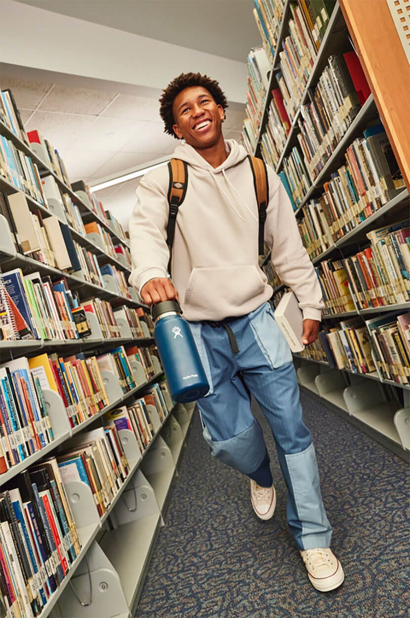 A young man smiling, walking through a library aisle, carrying a Hydr oFlask water bottle and a book, wearing a white hoodie, blue jeans, and a backpack.