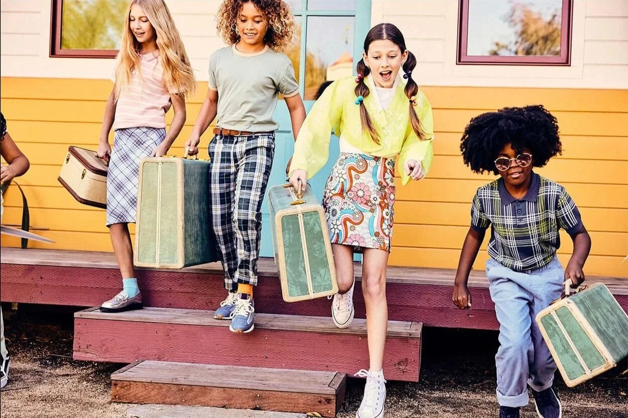 Four children, two girls and two boys, walking down steps outside a colorful house with blue and pink windows, carrying vintage suitcases and smiling.