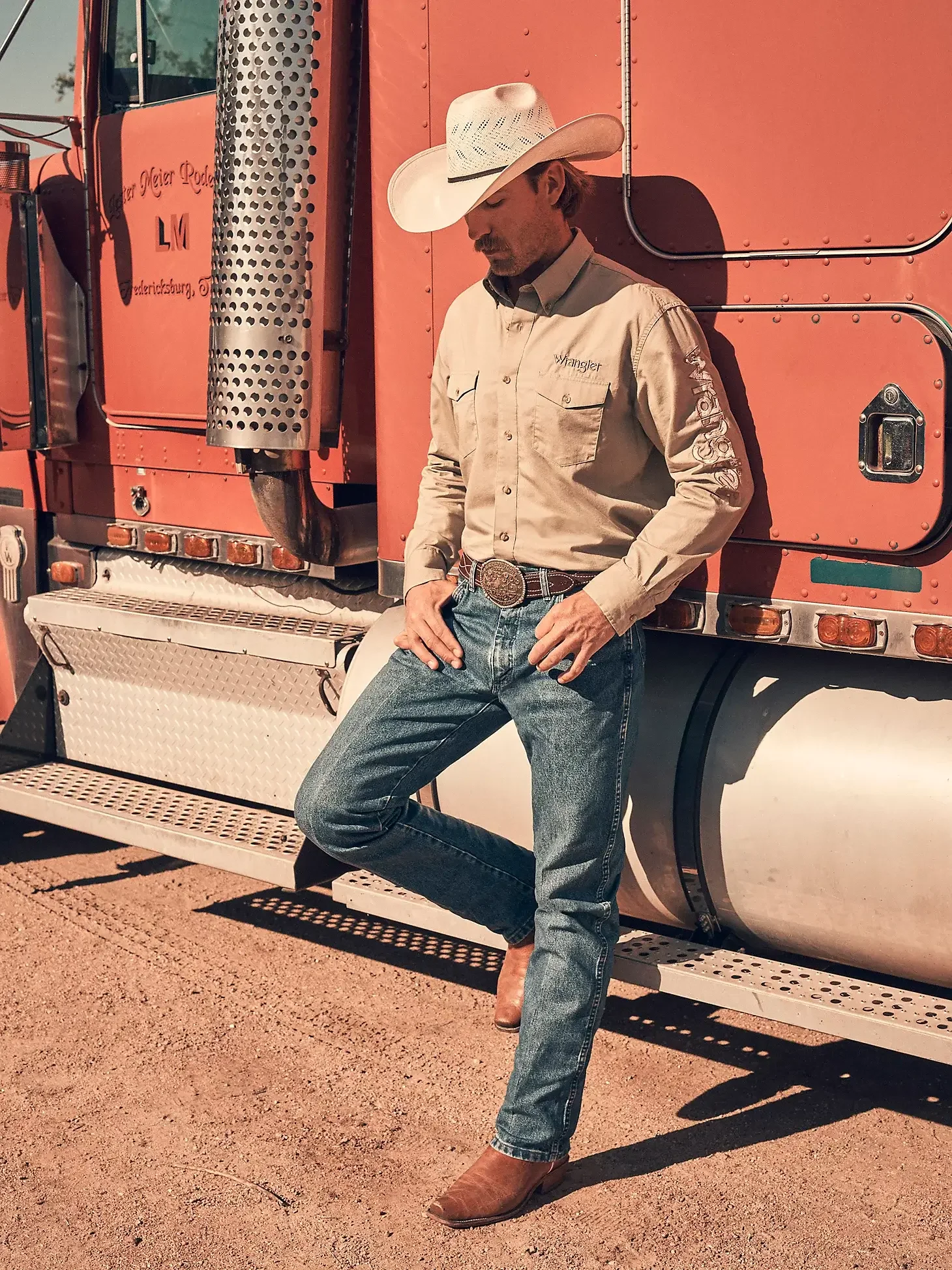 A man dressed in cowboy attire, including a cowboy hat, Wrangler shirt, jeans, brown cowboy boots, and a belt with a large buckle, leaning against a red semi-truck outdoors on dirt ground.