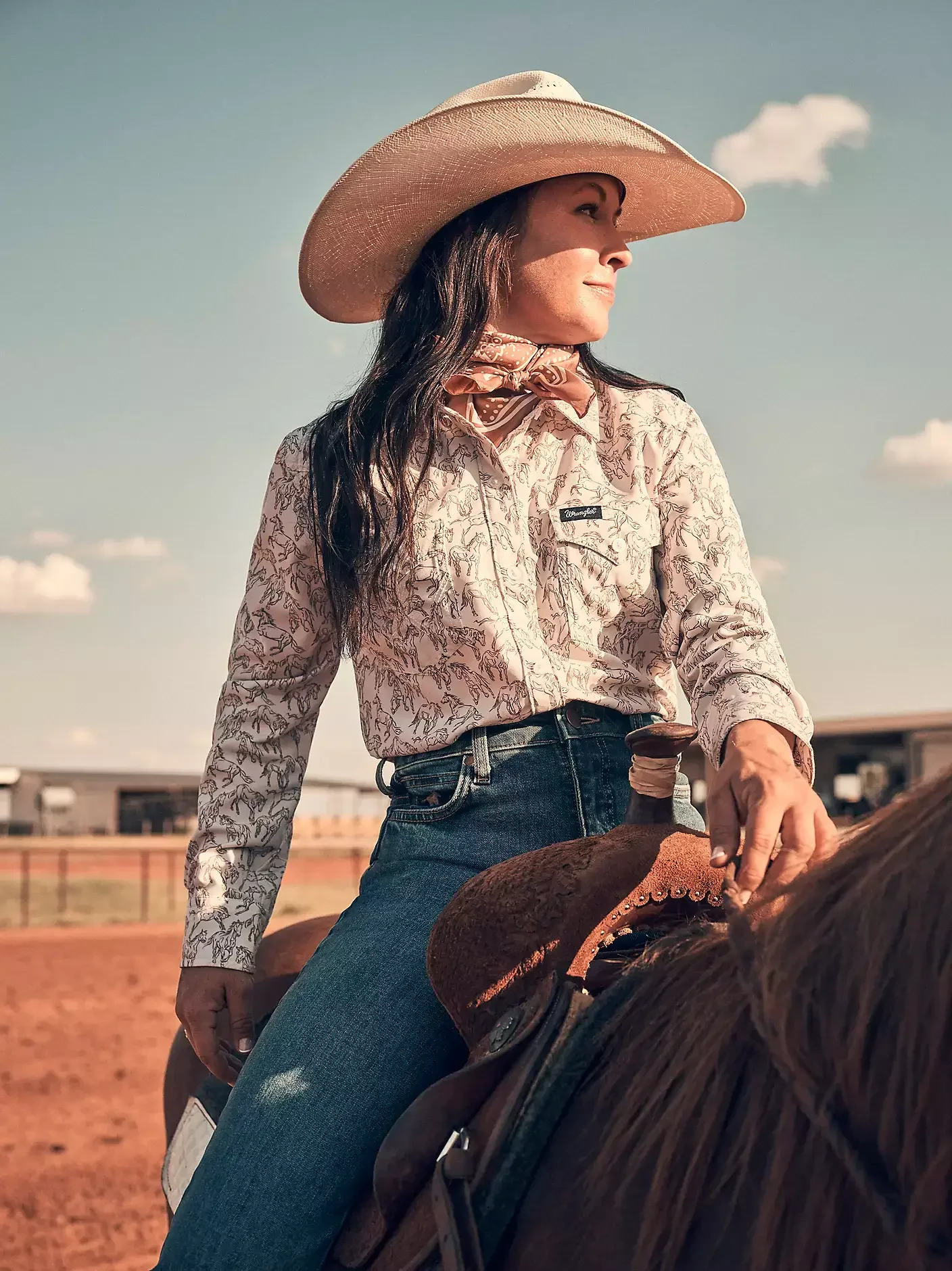 A woman wearing a cowboy hat, a patterned long-sleeve shirt, a bandana, and jeans, riding a horse in an outdoor arena on a sunny day.