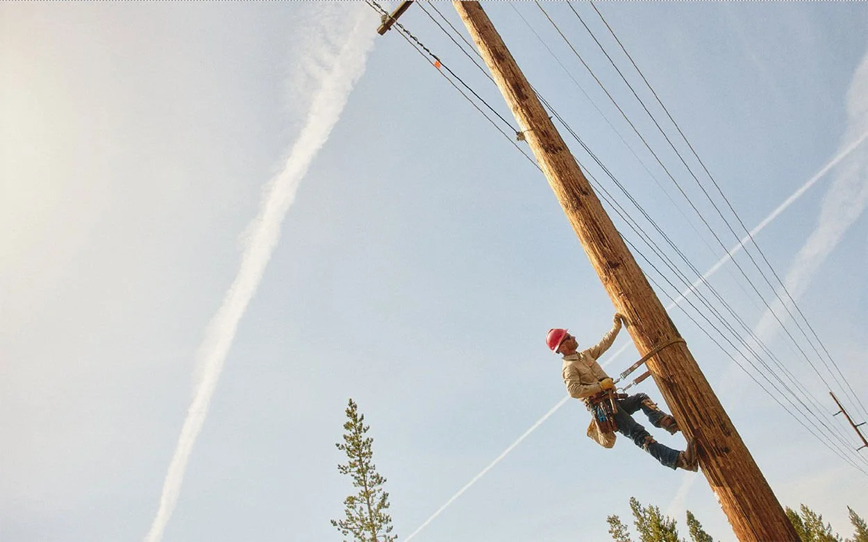 Worker climbing a wooden utility pole with Wranger clothing, safety gear and a red helmet, holding on to the pole while working on power lines against a clear sky with trees in the background.