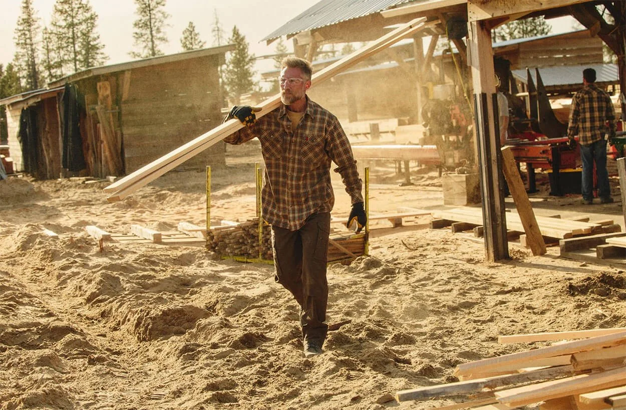 A man carrying a piece of wood in a construction yard wearing Wrangler pants and workwear with dirt ground, wooden structures, and trees in the background.