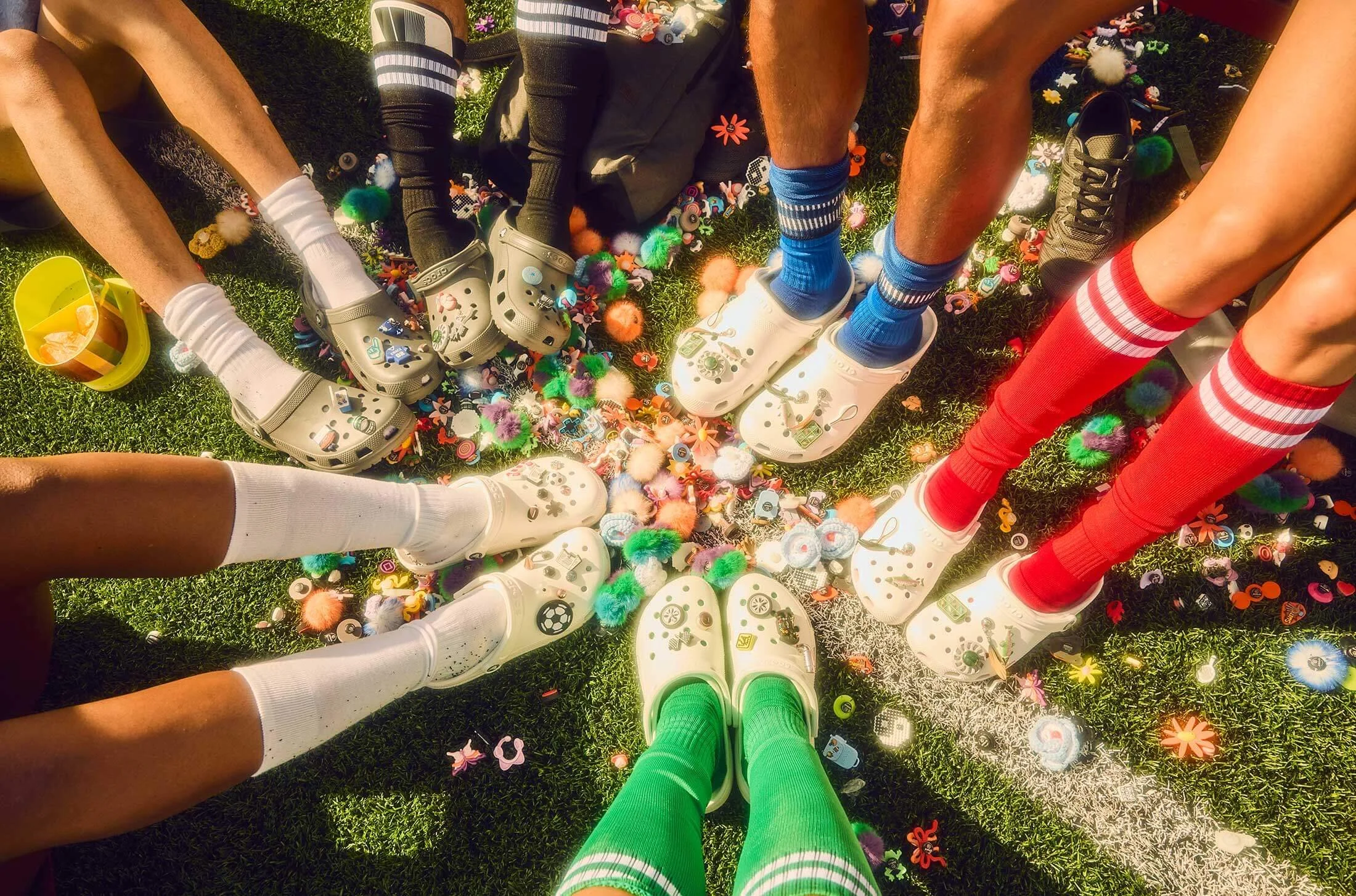 People with colorful socks and Crocs shoes sitting in a circle, surrounded by scattered small toys and craft supplies on grass.