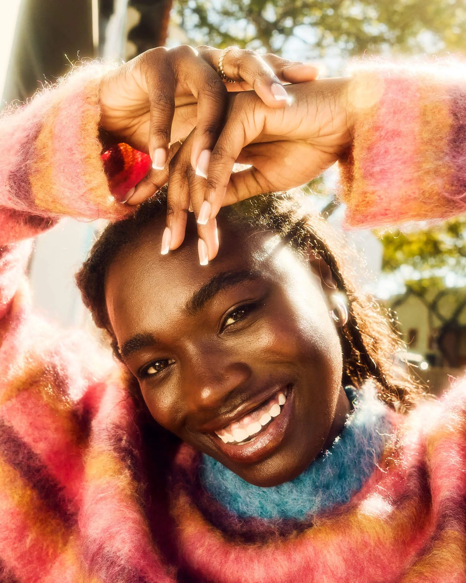 Close-up of a smiling woman with dark skin wearing a multicolored fuzzy sweater and a small hoop earring, holding her hands above her head with interlocked fingers, outdoors on a sunny day.