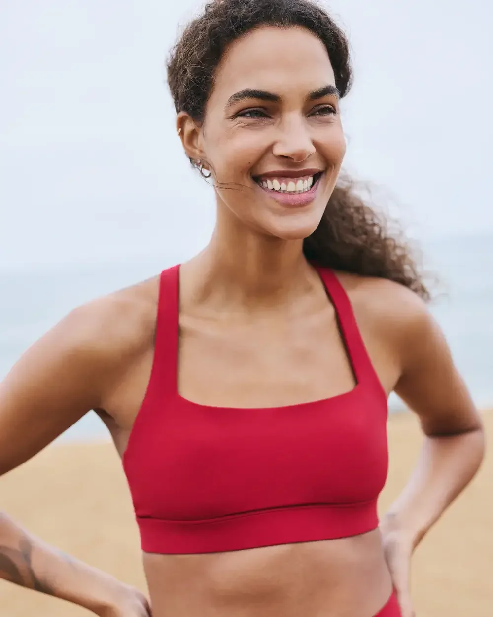 Smiling woman in red sports bra on a beach with a cloudy sky in the background.