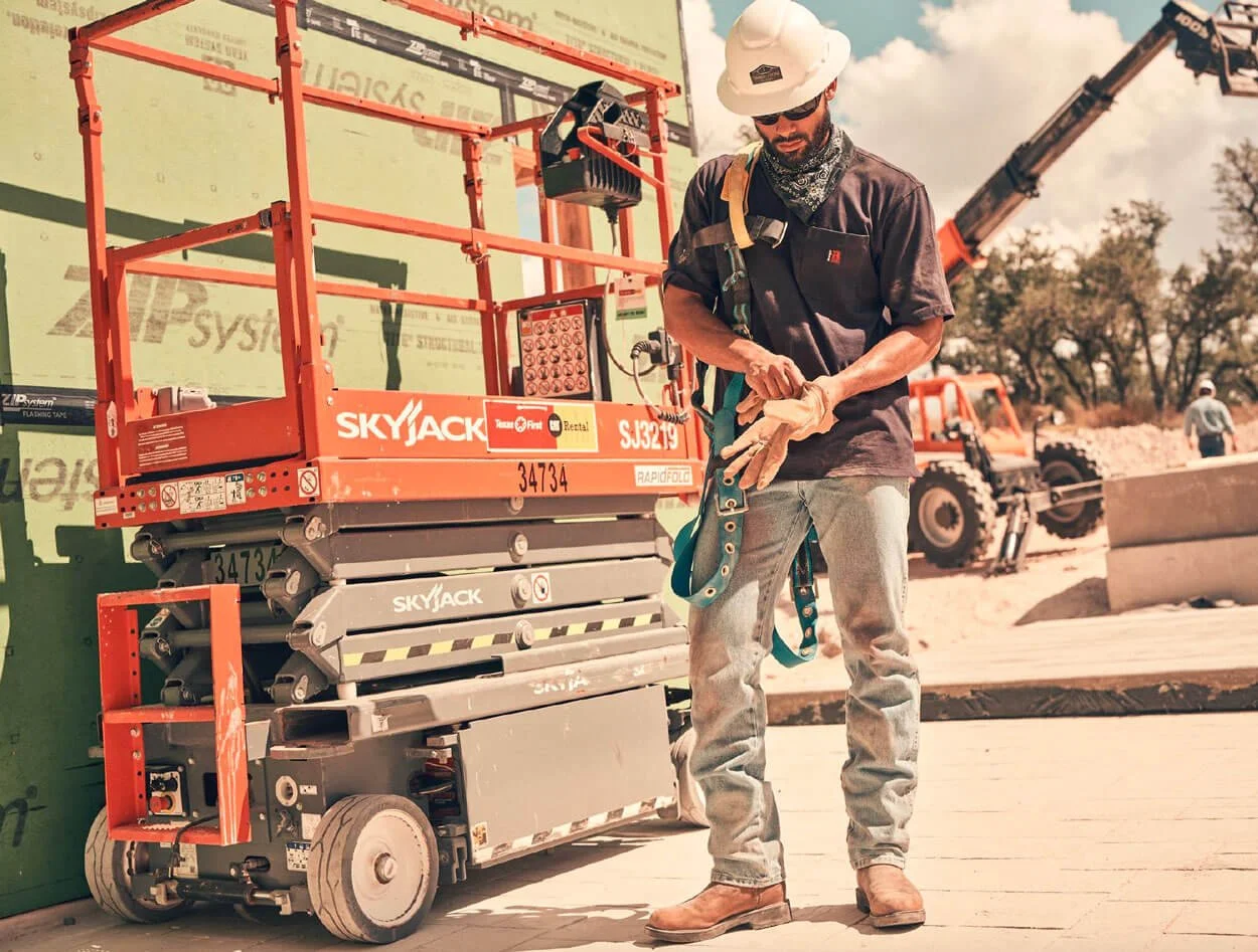 A construction worker with a hard hat and safety glasses adjusting his gloves at a construction site. Behind him is a large orange Skyjack lift machine and a crane in the background, with trees and a worker in the distance.