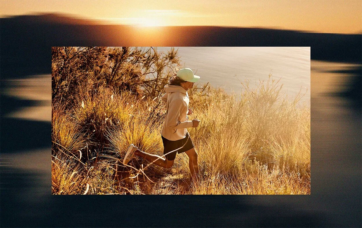 A woman jogging along a trail in a grassy area near water during sunset or sunrise.