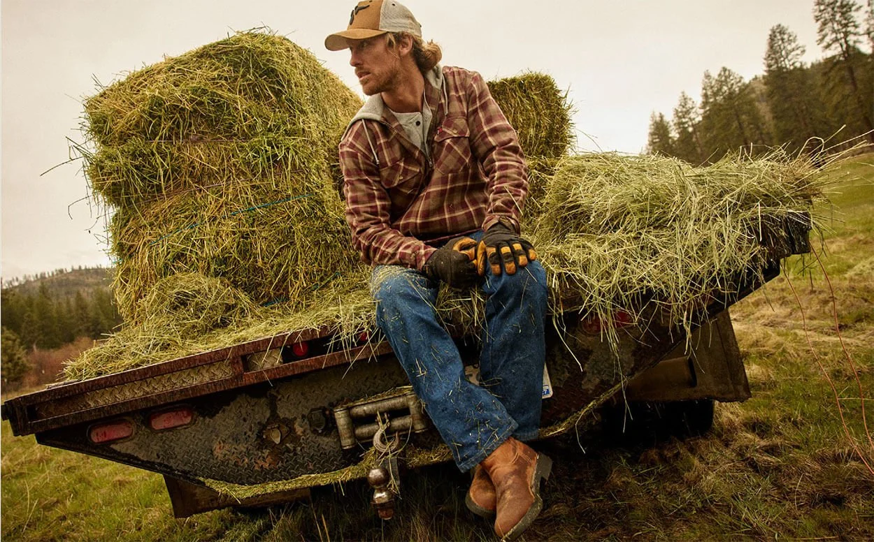 A man sitting on the side of a trailer filled with hay bales in a rural outdoor setting.