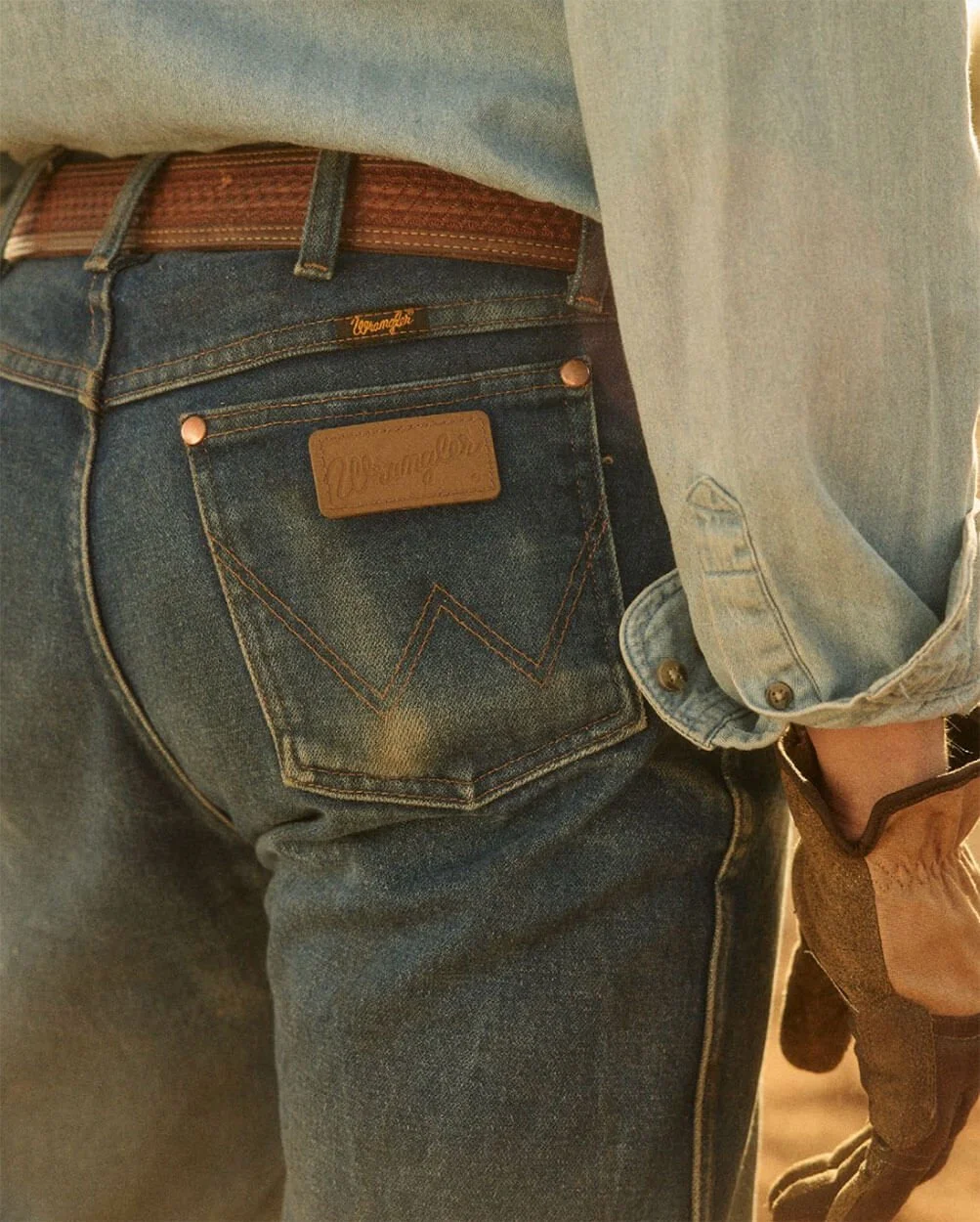 Close-up of a person wearing blue denim jeans, a light-colored shirt, and a brown belt. The jeans have a leather patch on the back pocket with the brand name 'Wrangler' and orange stitching.