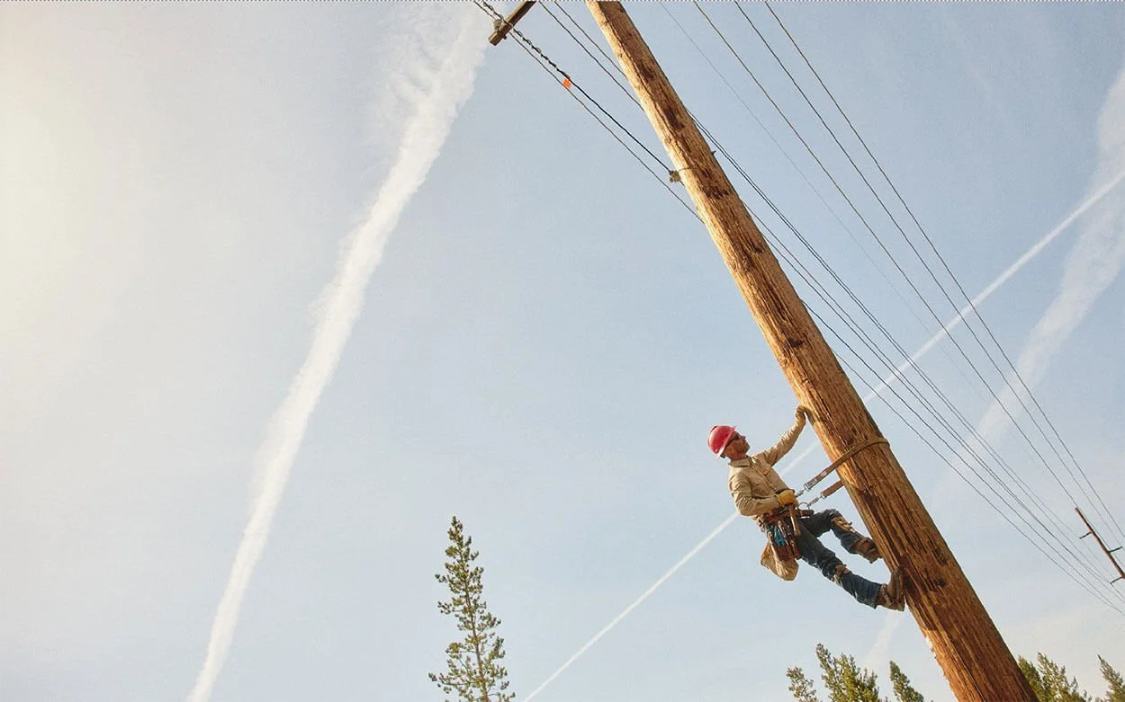 A utility worker wearing a red helmet and safety harness is climbing a wooden utility pole with power lines, surrounded by trees and a blue sky with clouds and contrails.