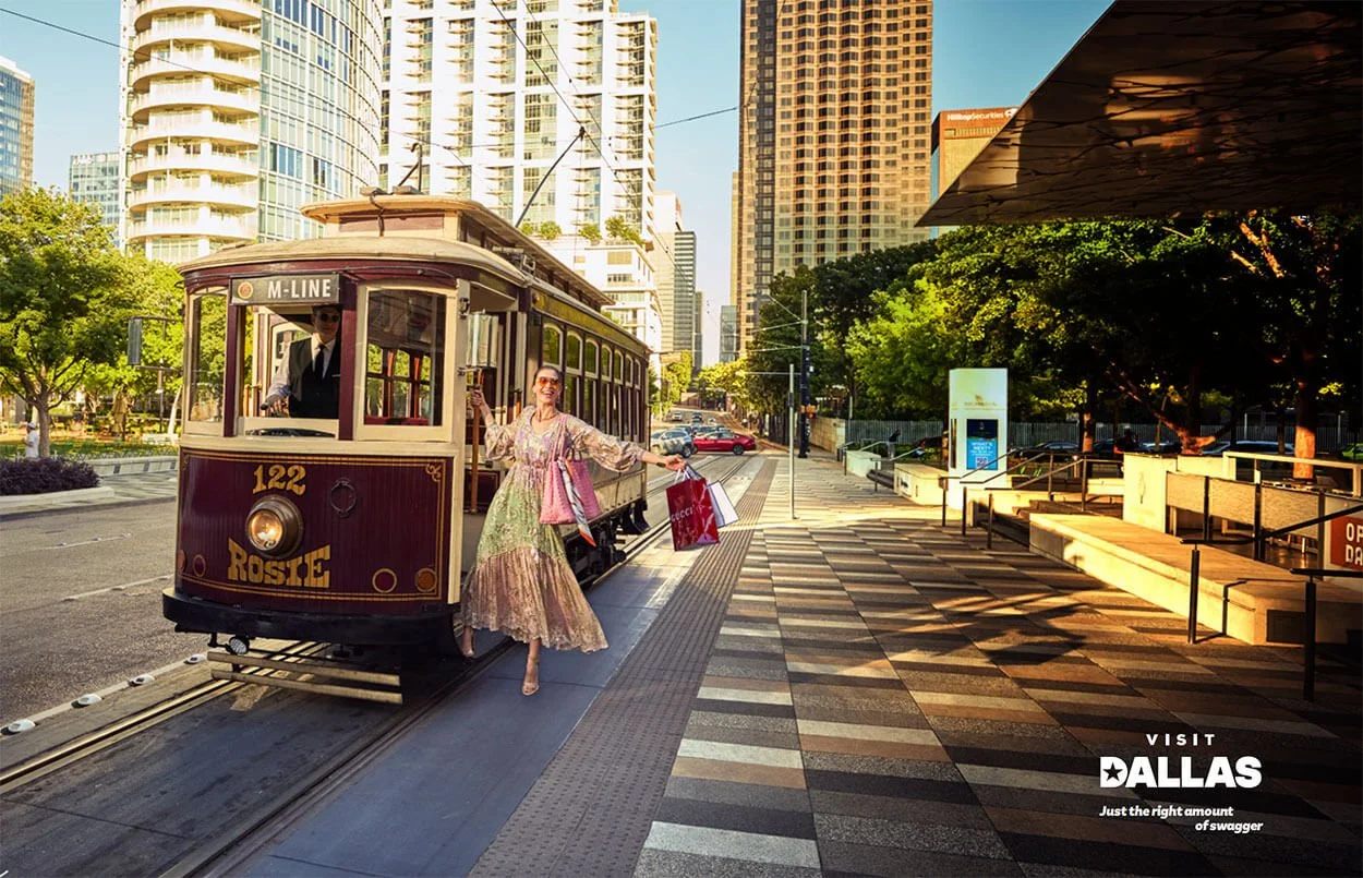 A woman in a colorful dress carrying shopping bags steps off a vintage tram labeled 'Rosie' and 'M-LINE' in an urban downtown area with high-rise buildings, trees, and a sunny sky.