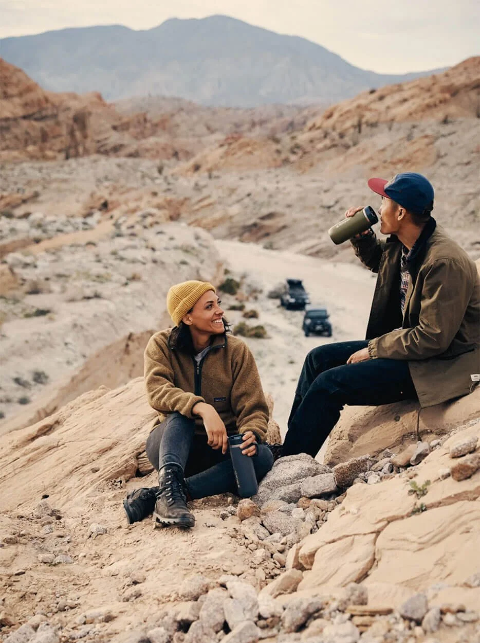 A man and woman sitting on rocky desert terrain, having a conversation; the woman is smiling and holding a water bottle, while the man is drinking from a thermos. Both are dressed in outdoor gear, with mountains and off-road vehicles in the backgroun