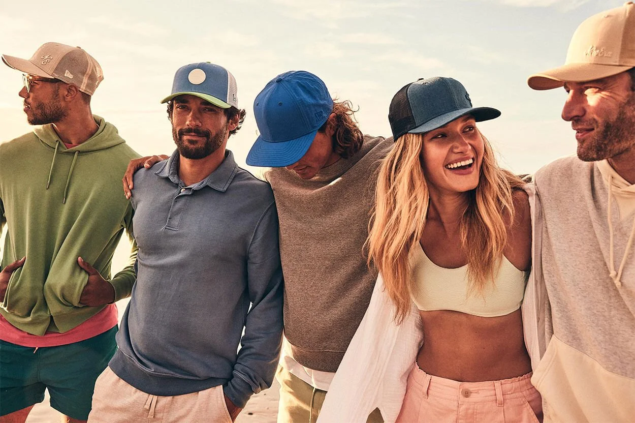 Group of five young adults standing together outside during sunset, smiling and enjoying each other's company, dressed casually in hoodies, caps, and shorts.