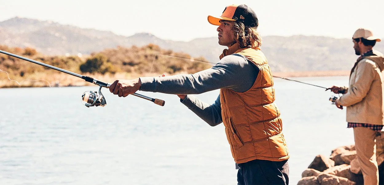 Two men fishing by a lake with mountains in the background, one in the foreground wearing an orange vest and baseball cap, holding a fishing rod.