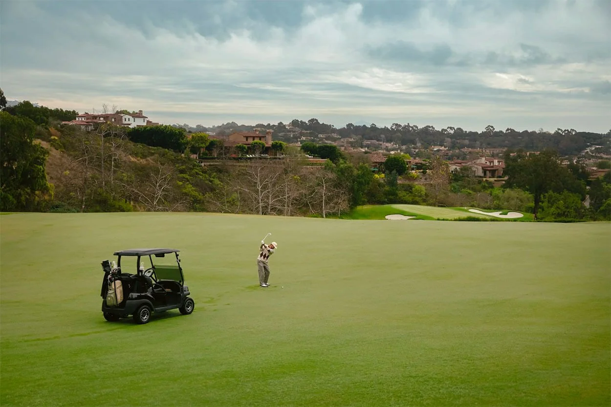 A golfer swinging a club on a golf course with a golf cart nearby and a scenic background of trees, houses, and a cloudy sky.