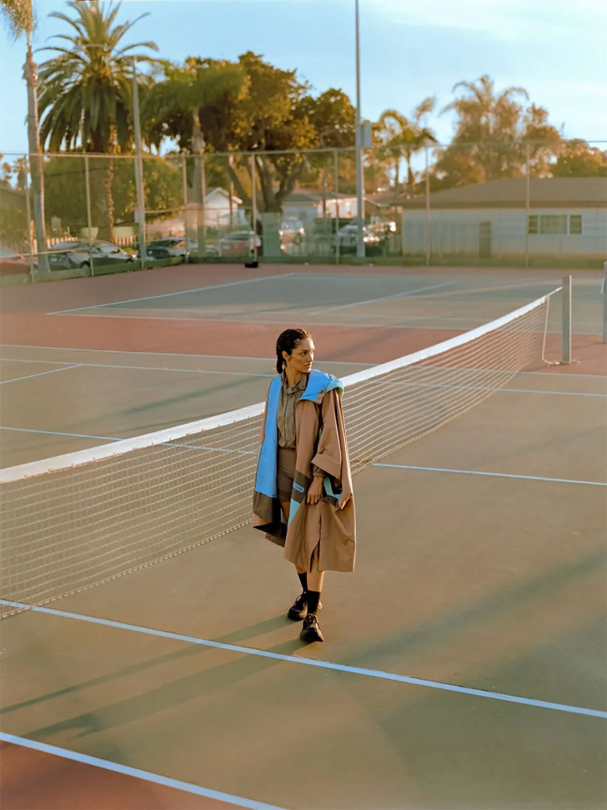 A woman standing on a tennis court at sunset, wearing a tan oversized coat with blue accents, black boots, and a beige outfit underneath.