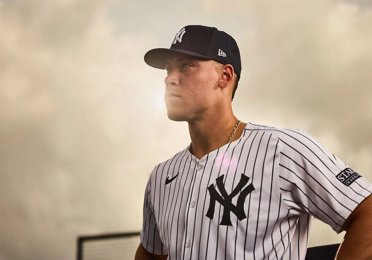Aaron Judge wearing a New Era New York Yankees cap, standing outdoors with a cloudy sky in the background.