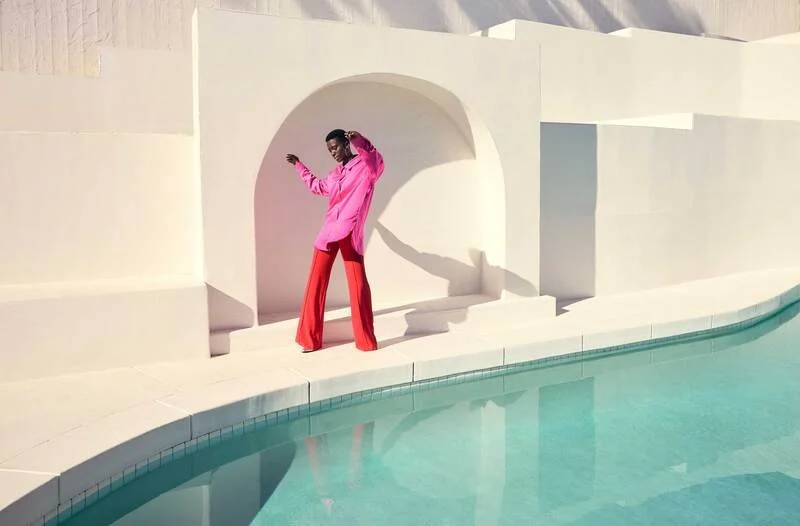 A person in a pink top and red pants standing by a pool, casting a shadow on a white wall with architectural arches.