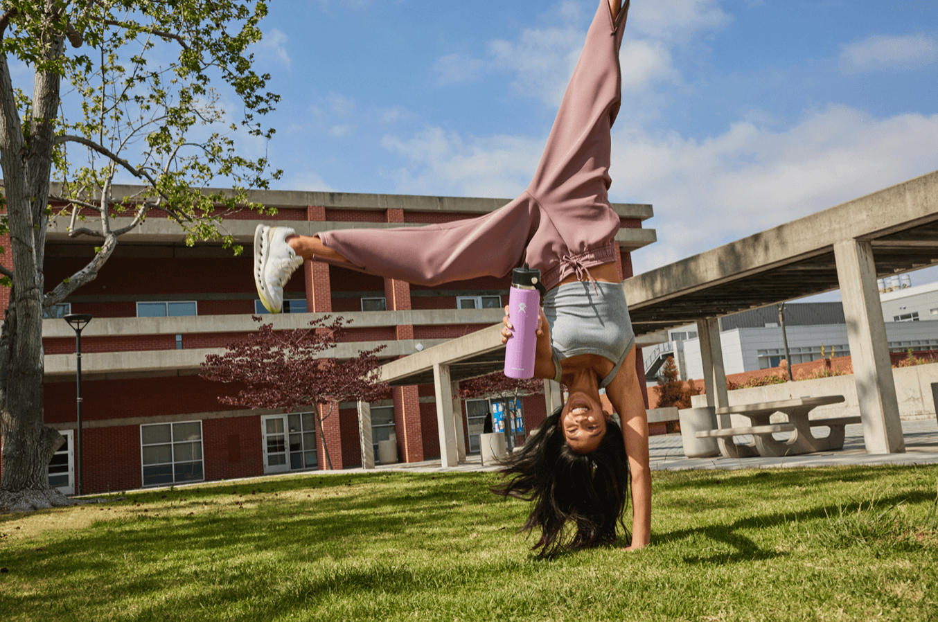 A woman doing a handstand on grass in an outdoor park or courtyard, holding a pink Hydro Flask water bottle, with trees and modern buildings in the background, under a partly cloudy sky.