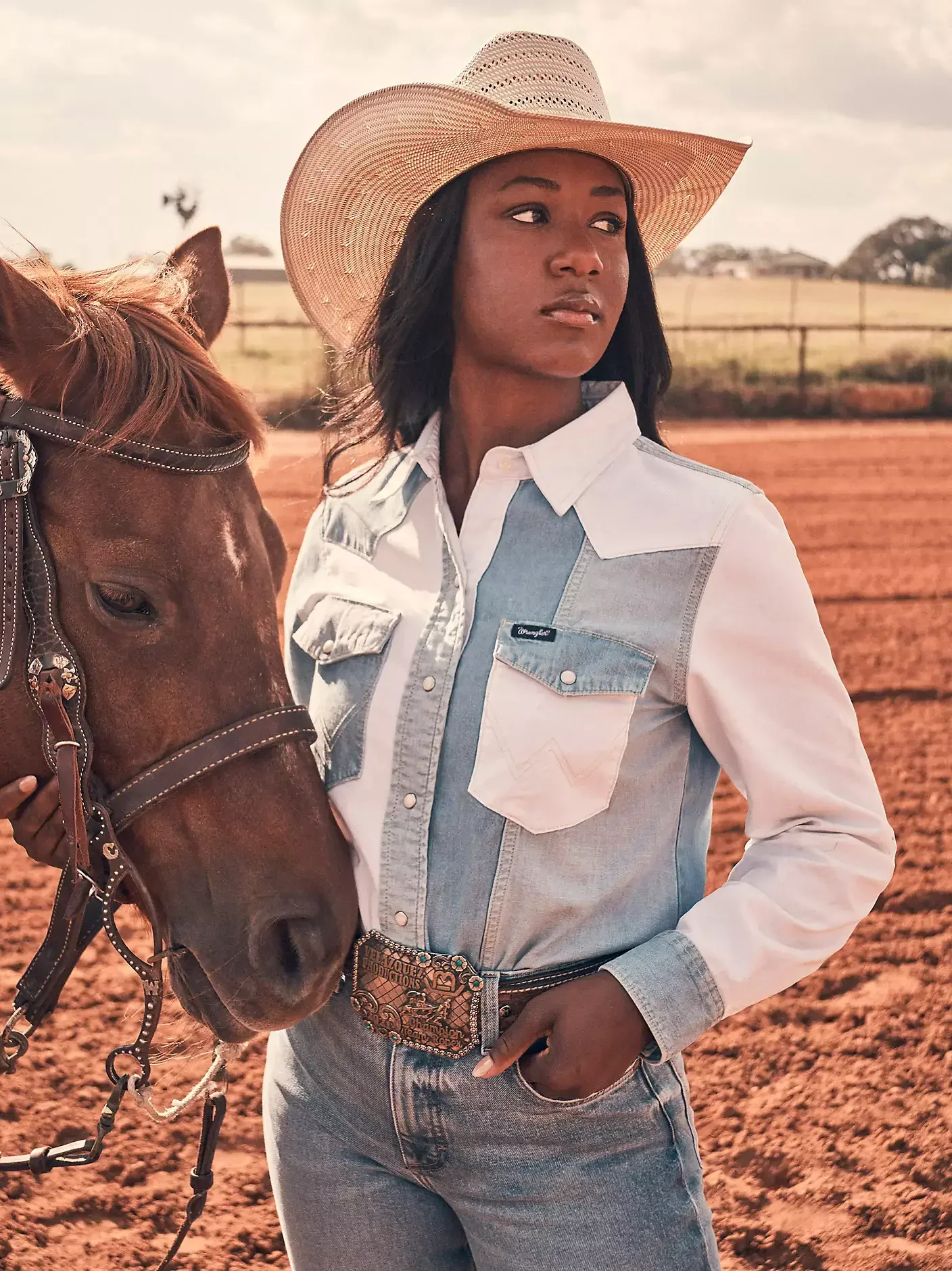 A woman standing next to a brown horse on a ranch, wearing a cowboy hat, a denim and white button-up shirt, and jeans with a large belt buckle.