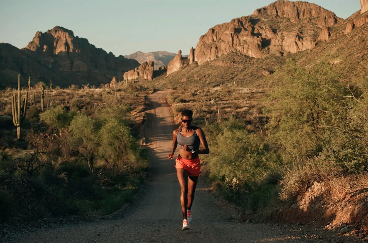 A woman running on a dirt trail in a desert landscape with cacti and rugged mountains in the background during sunset.