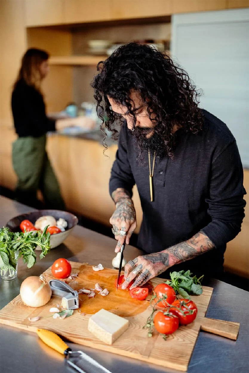 A man with tattoos and curly black hair slicing a tomato on a wooden cutting board in a kitchen, with fresh vegetables and herbs around him and another person in the background.