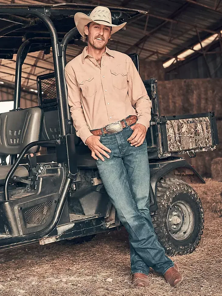 A man wearing a cowboy hat, beige button-up shirt, jeans, and cowboy boots leaning against a utility vehicle inside a barn.