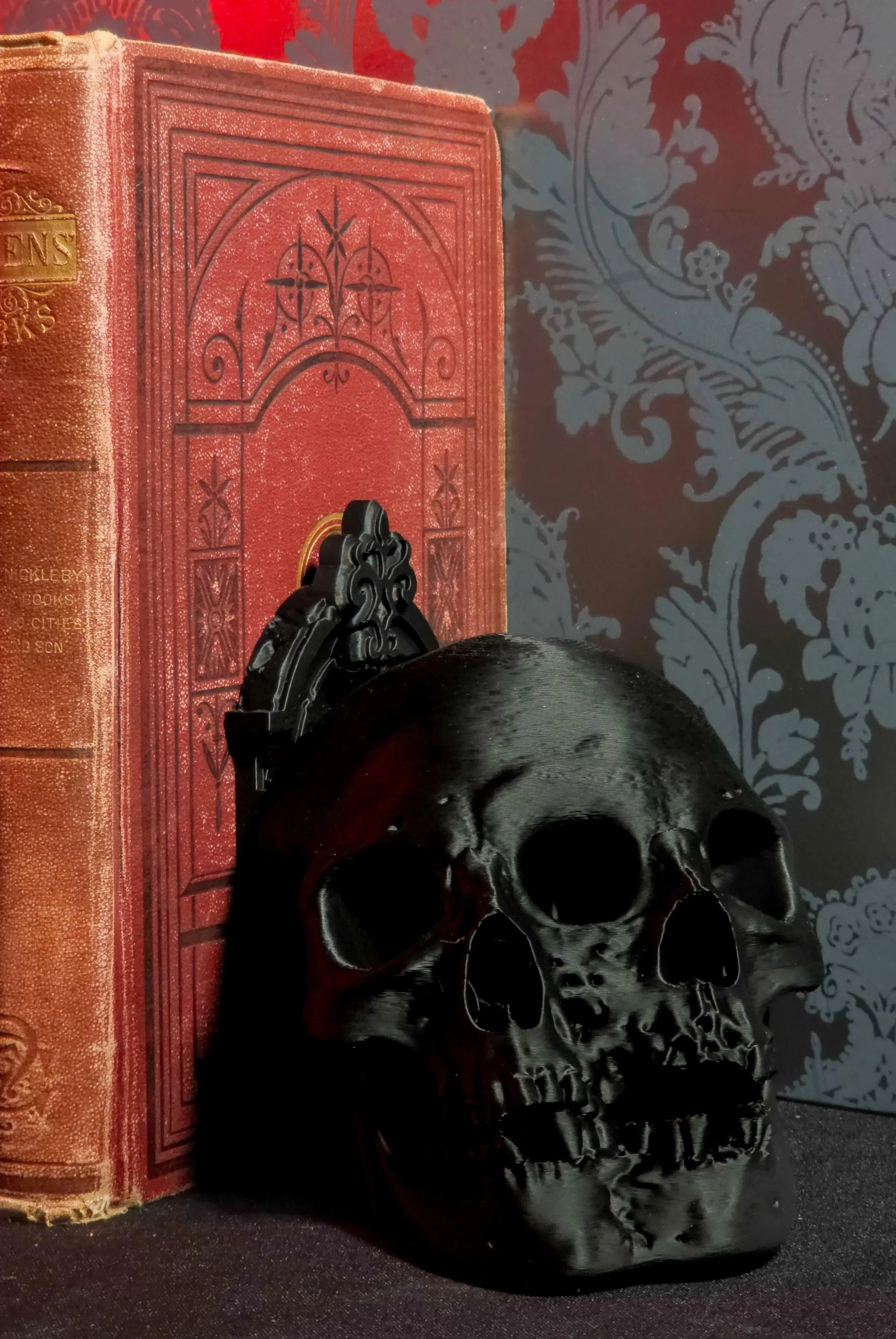 A black skull sculpture placed on a dark surface next to an old red book with ornate gold details on the spine, against a background with decorative gray floral pattern.
