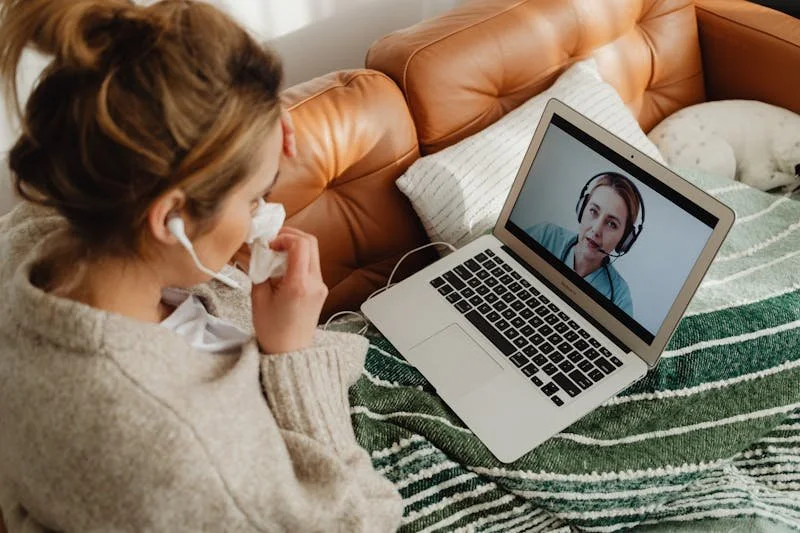 Woman on a couch with a laptop in her lap. She is participating in online therapy with another woman. Both of them are wearing headphones.