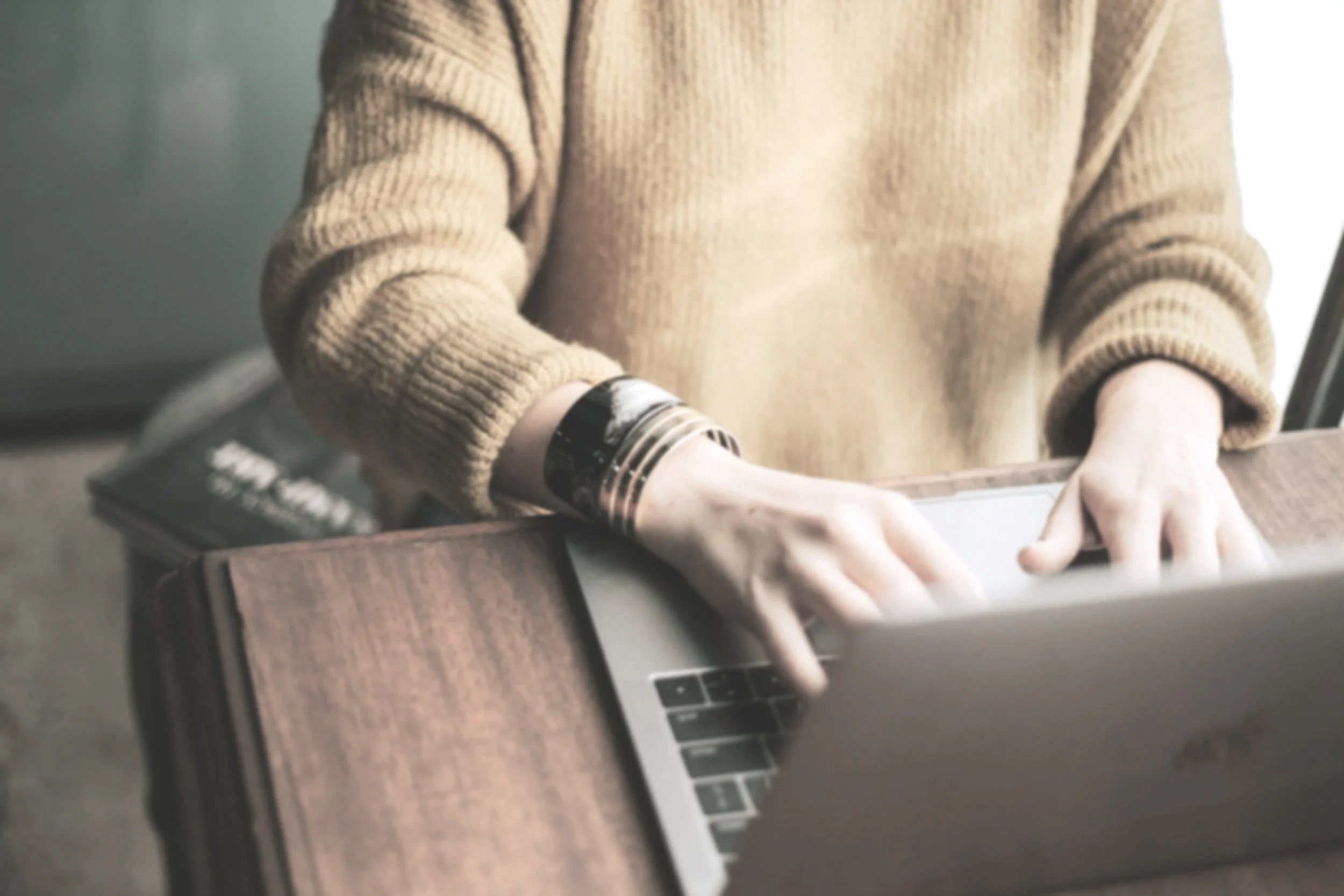 The torso and hands of a person in a yellow sweater and wearing gold bracelets can be seen resting on a brown wooden table and typing on a laptop computer.